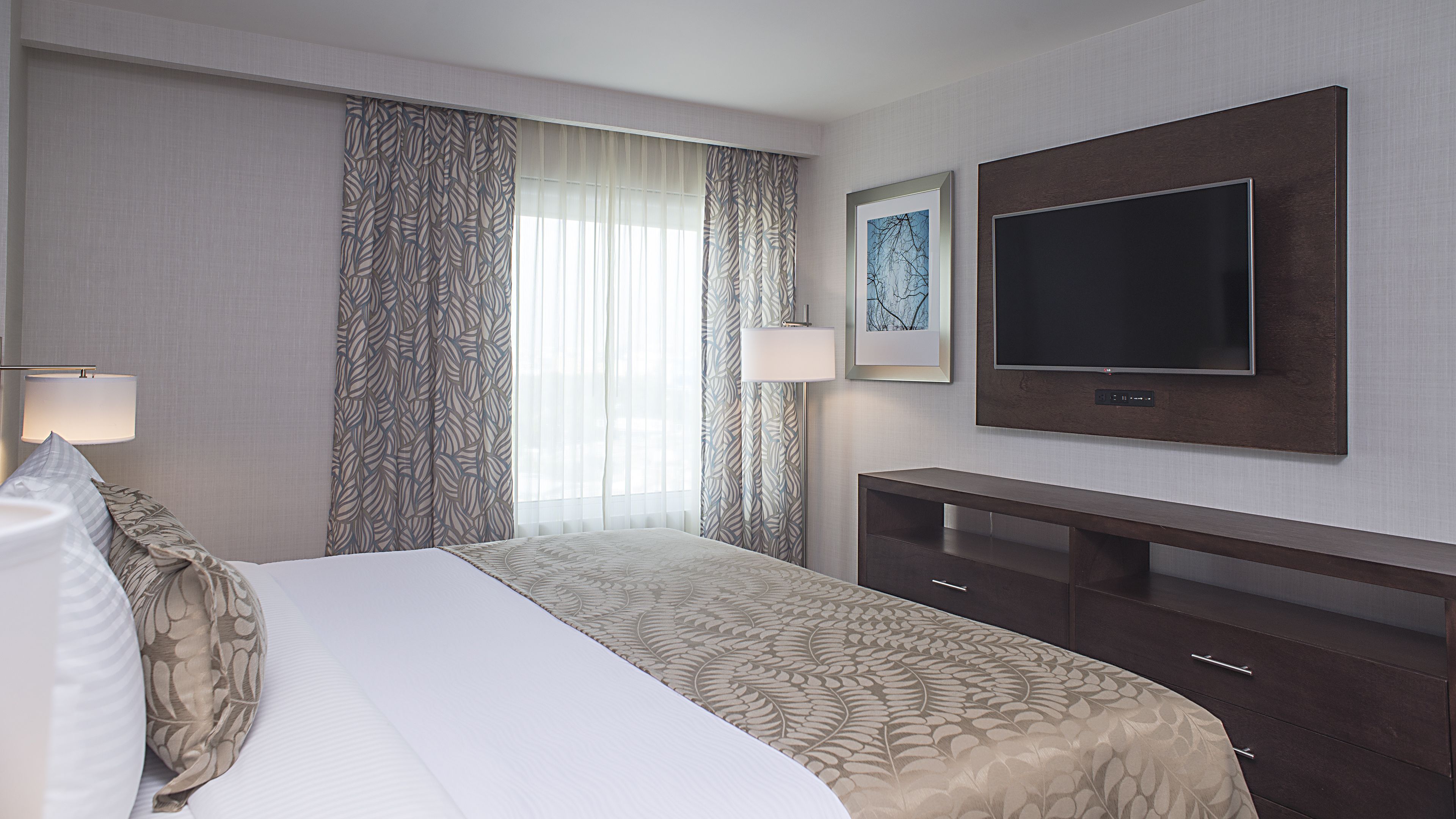 A hotel room with a bed, a TV mounted on the wall, a desk, and a lamp.