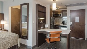 Una habitación de hotel pequeña con cama, escritorio, silla, kitchenette y espejo.