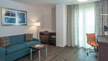 Habitación de hotel con sofá, una mesita, escritorio con silla, lámpara y ventana con cortinas.
