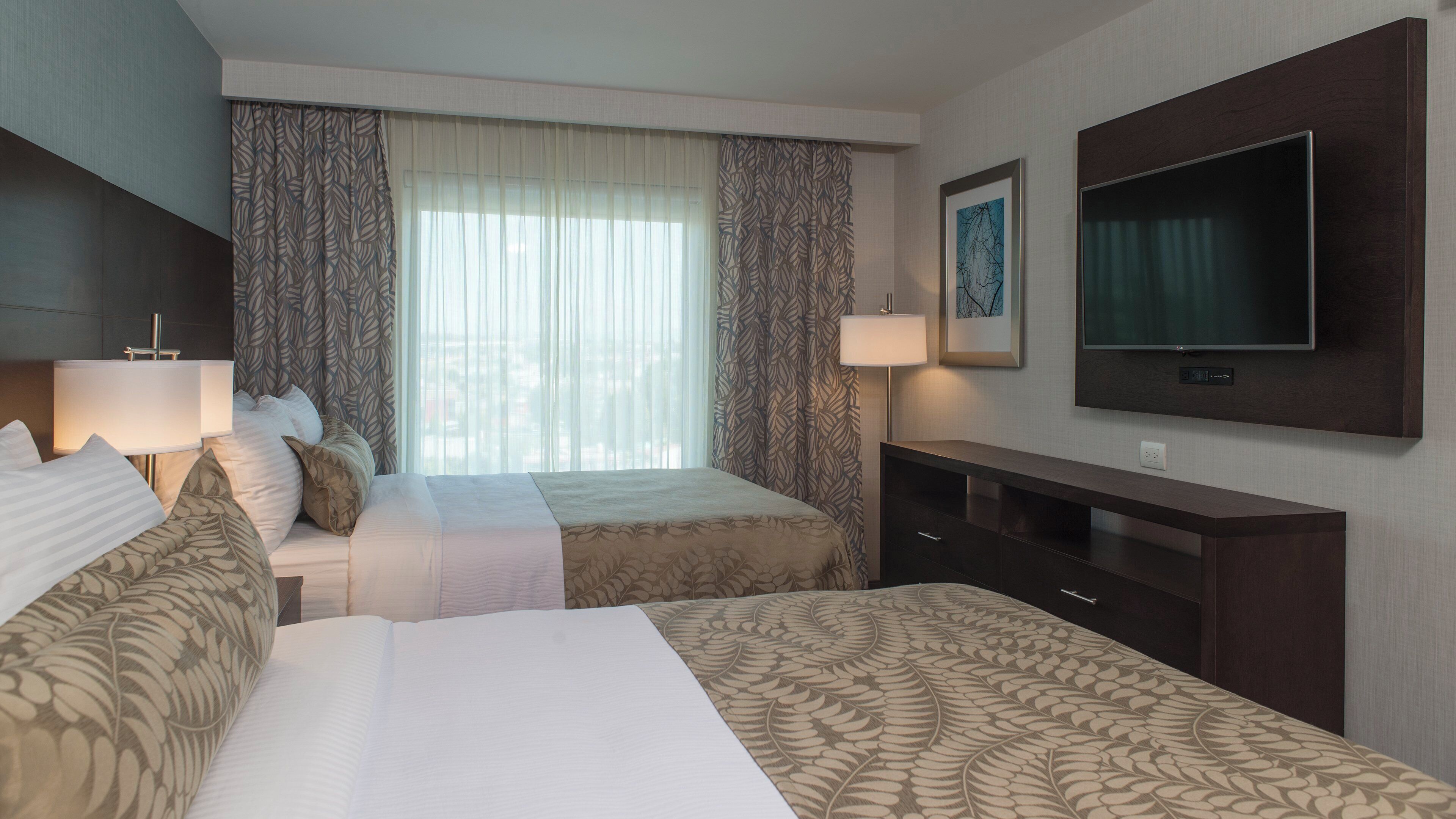 Habitación de hotel con dos camas, televisión, escritorio y ventana con cortinas.
