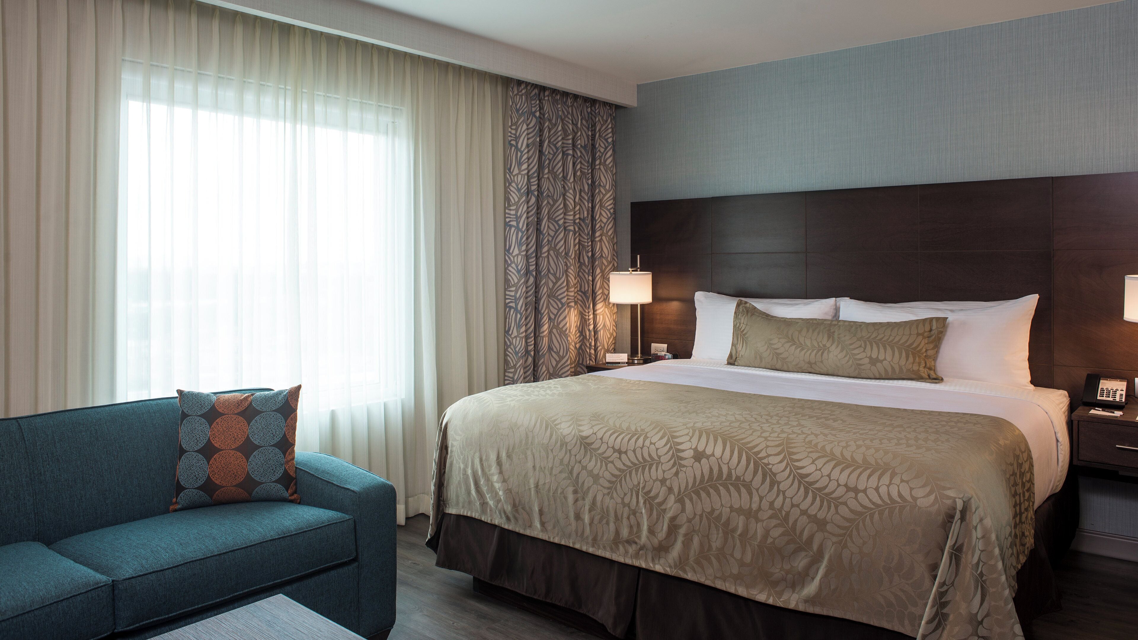 Una habitación de hotel con una cama, una mesita de noche, una lámpara, un sofá y una ventana con cortinas.