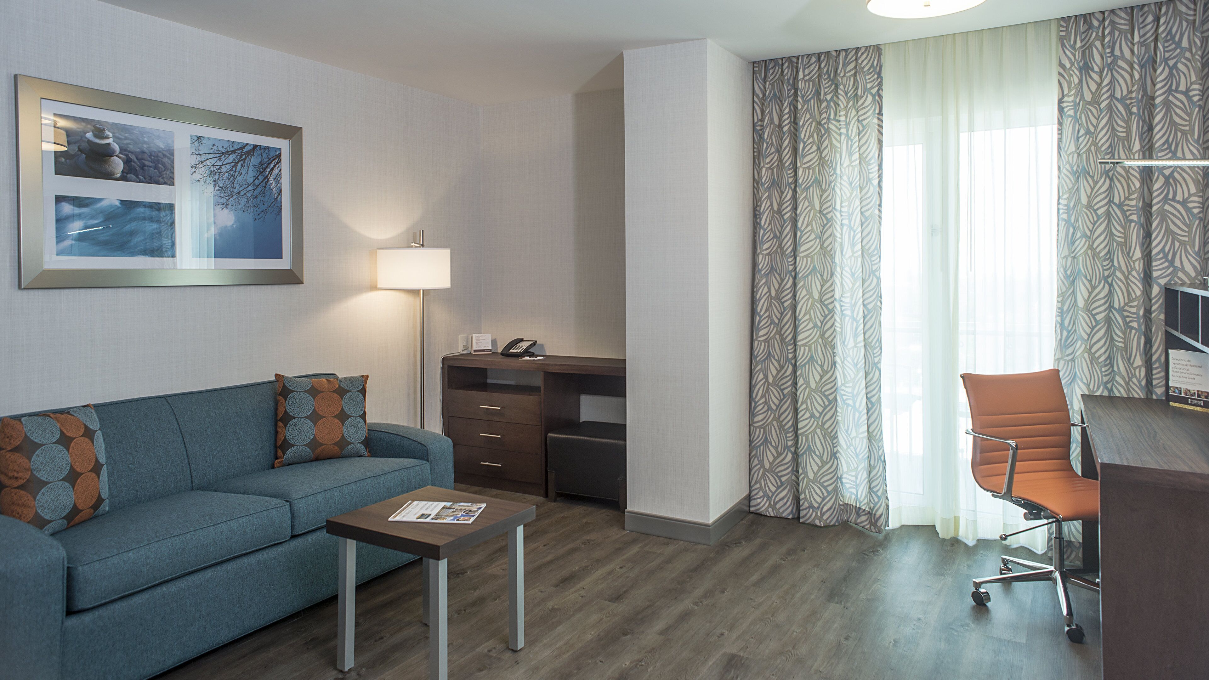 Una habitación de hotel moderna con un sofá azul, una mesita de madera, un escritorio con una lámpara y una cortina estampada.