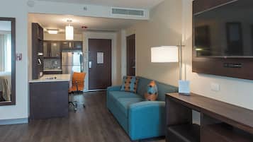A hotel room with a kitchenette, a sofa with patterned cushions, a desk, and a flat-screen TV.