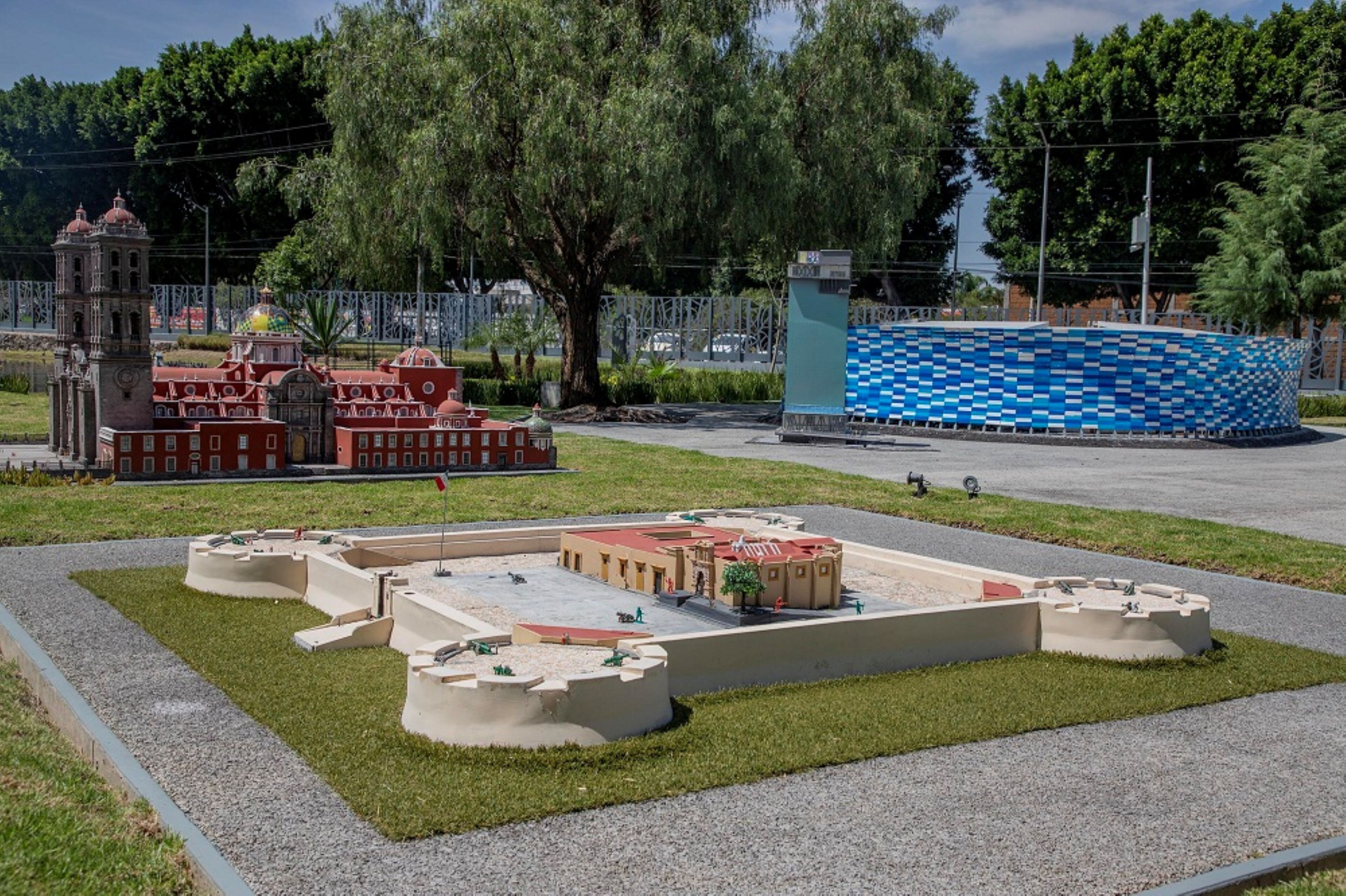 A model of a building complex with a central courtyard, surrounded by a fence, and a large blue mosaic structure in the background.