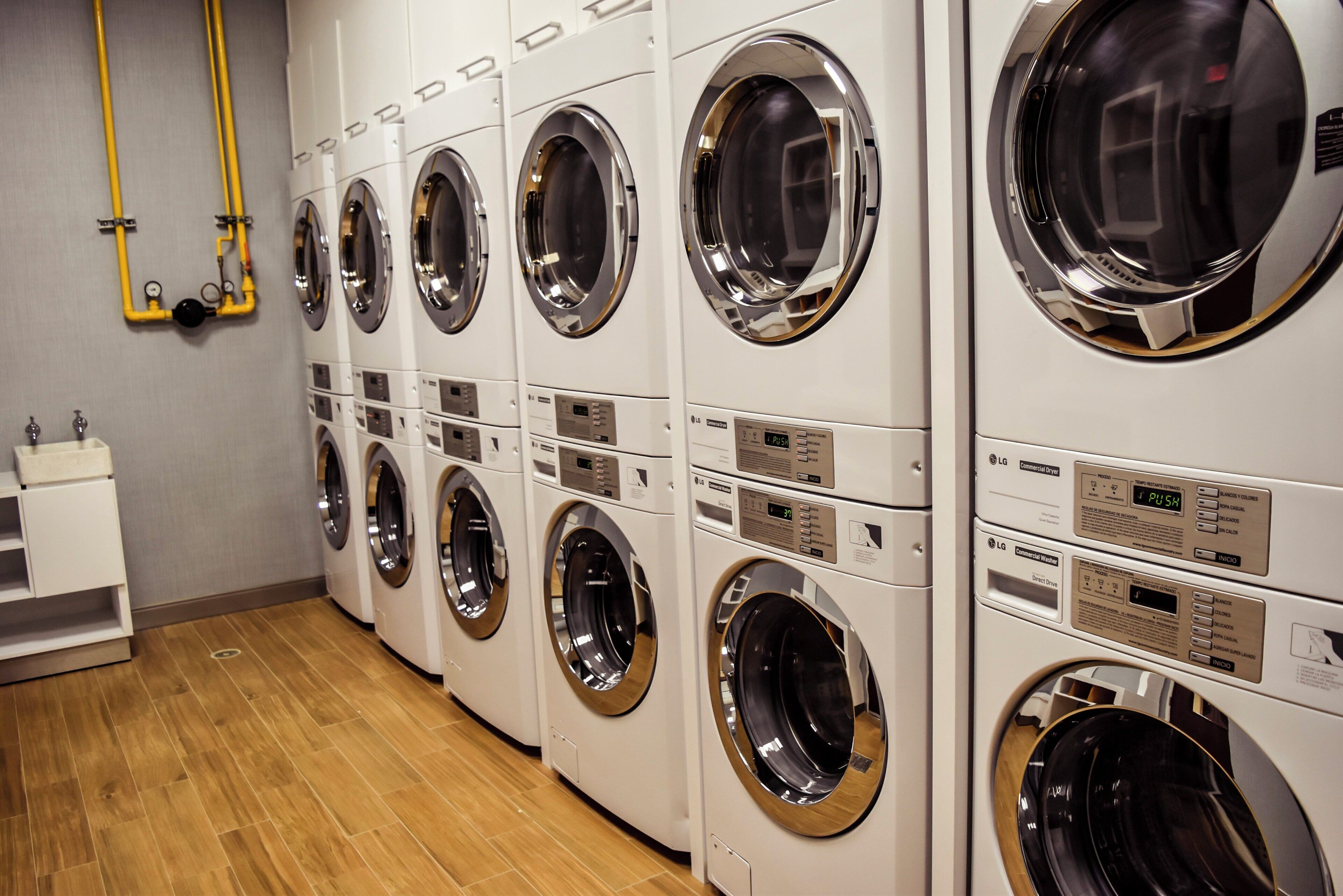 A row of white commercial washers with control panels, a wooden floor, and a small white shelf with a sink.