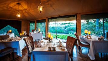 Un comedor interior con mesas dispuestas para dos personas, ventanas amplias y lámparas colgantes.