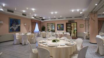 Une salle de banquet avec des tables rondes dressées pour le repas, des chaises blanches et des luminaires au plafond.