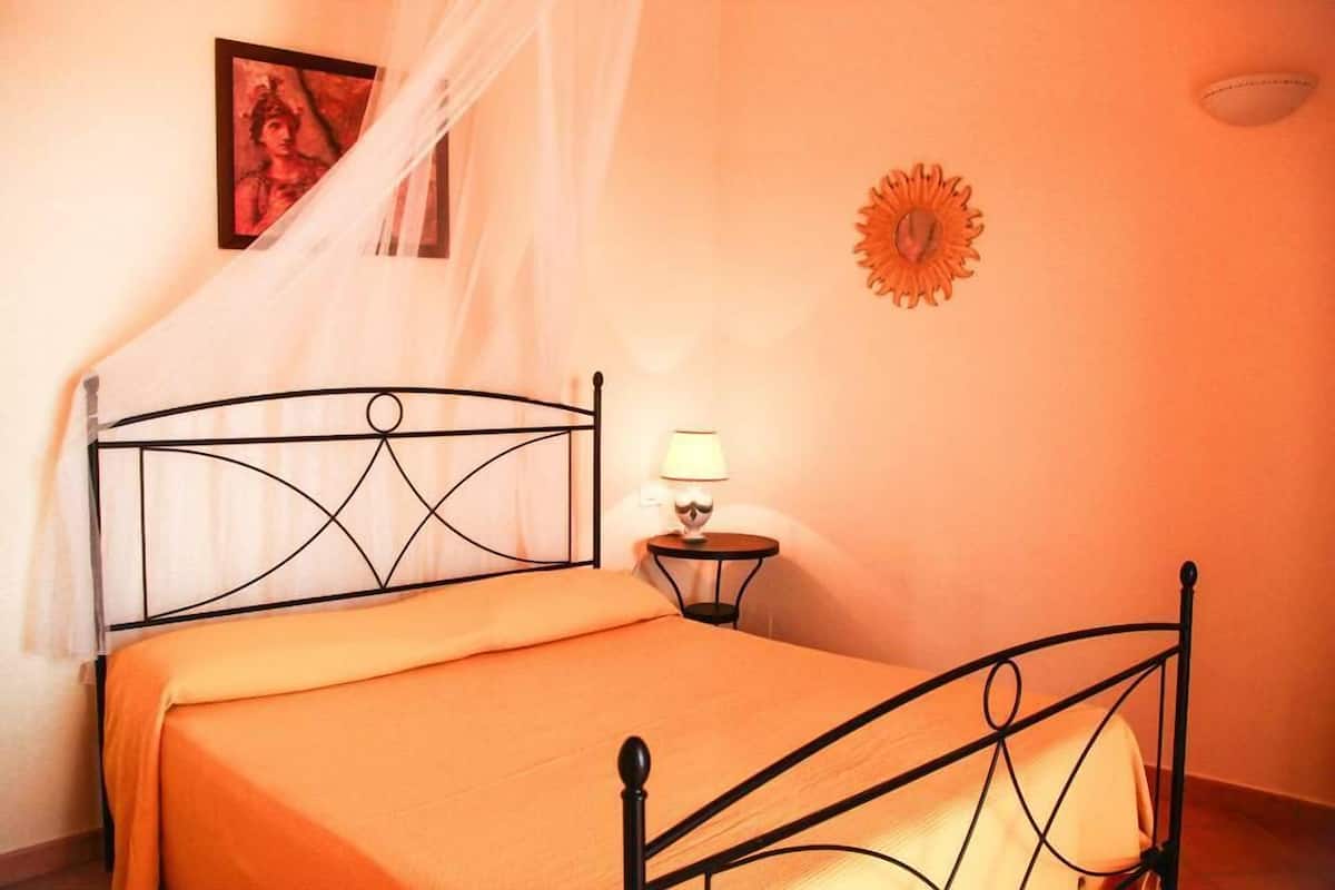 A room with a bed, a nightstand, a lamp, a picture on the wall, and a decorative sun-like object.