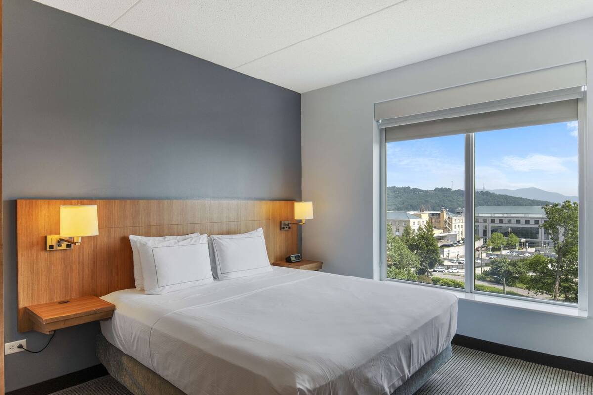 Ein Hotelzimmer mit einem großen Bett, zwei Nachttischen mit Lampen und Blick auf eine Stadtlandschaft durch ein großes Fenster.