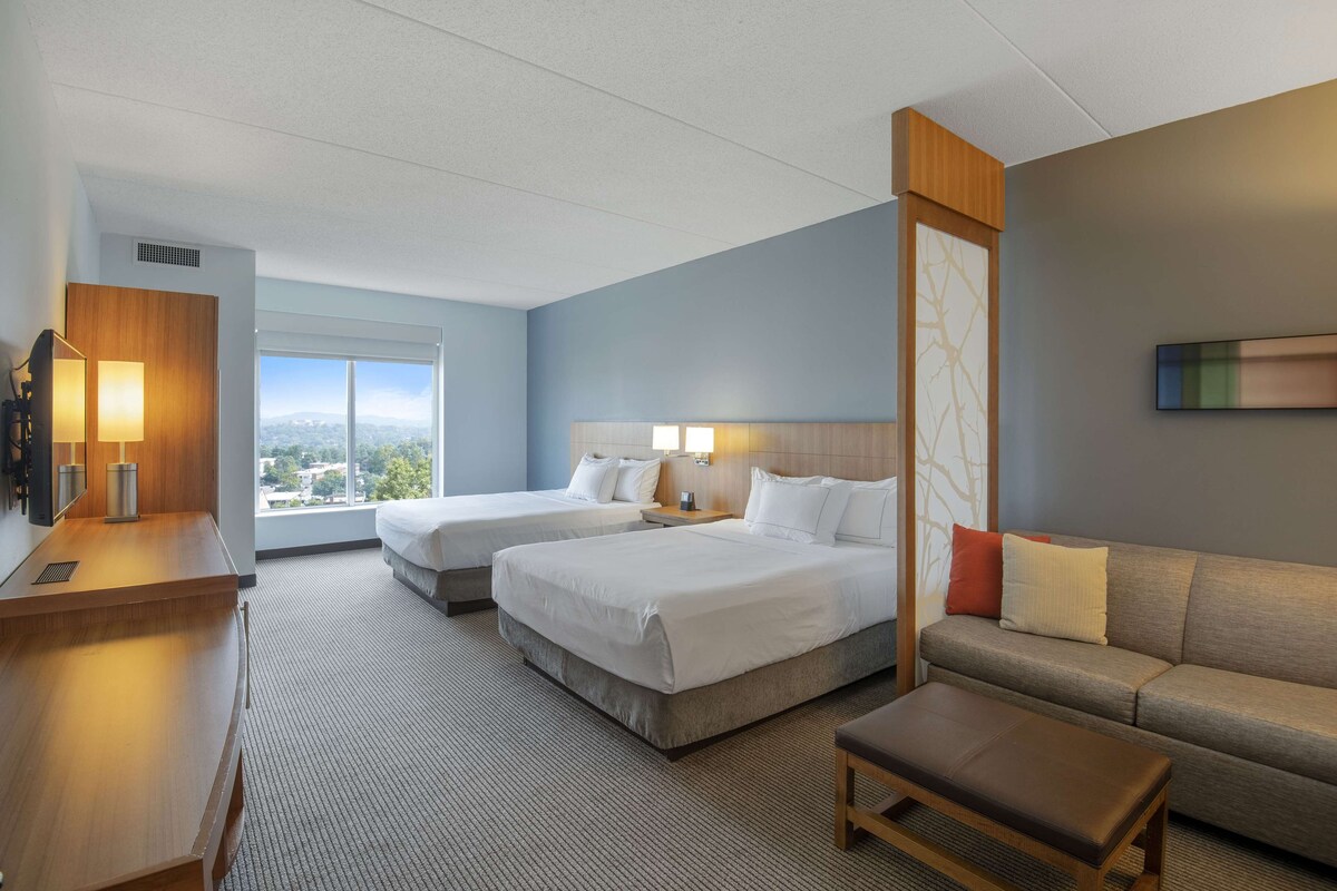A hotel room with two beds, a sofa, a desk, and a large window offering a city view.
