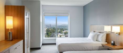 Habitación de hotel con dos camas, un tocador de madera, un escritorio con lámpara y un ventanal con vistas a la ciudad.