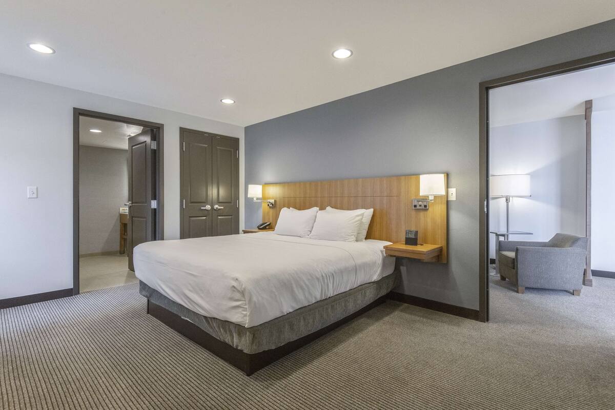 Ein modernes Hotelzimmer mit einem großen Bett, einem Holzkopfbrett, Nachttischlampen, einem kleinen Tisch und einem Sessel.