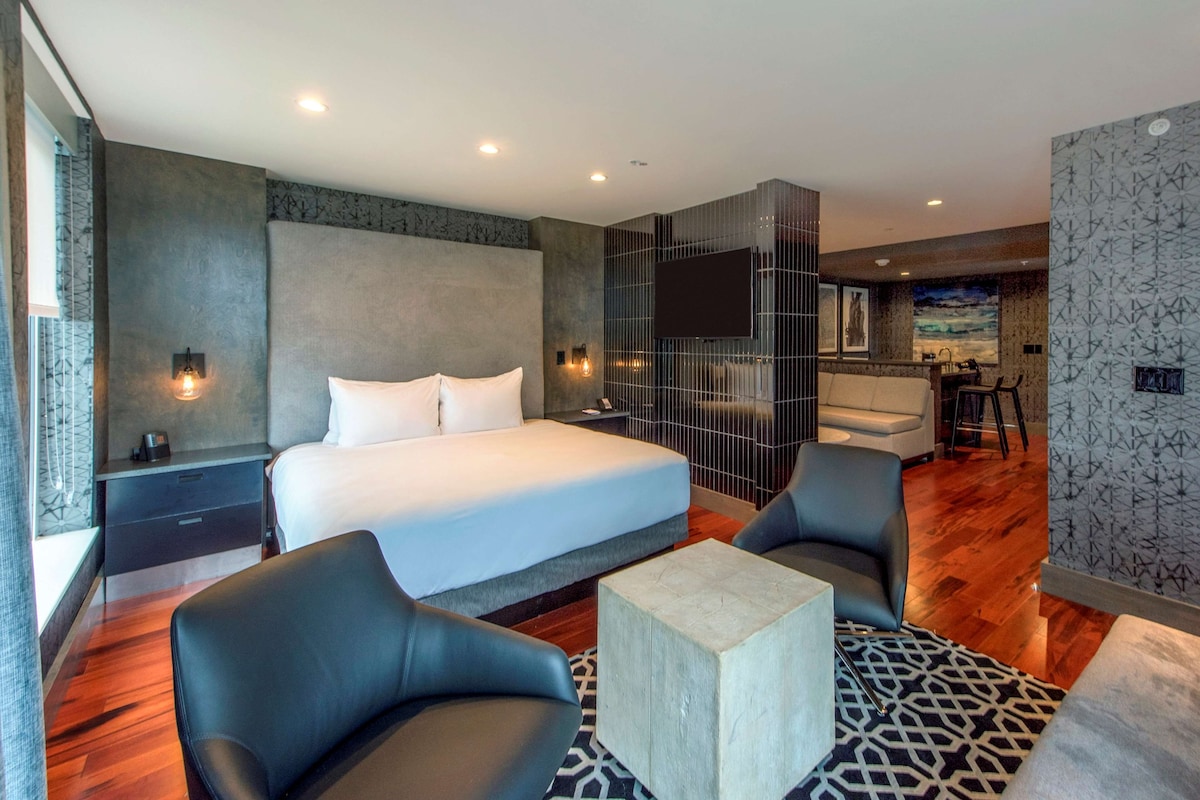 A modern hotel room with a large bed, two armchairs, a small coffee table, and a TV mounted on the wall.