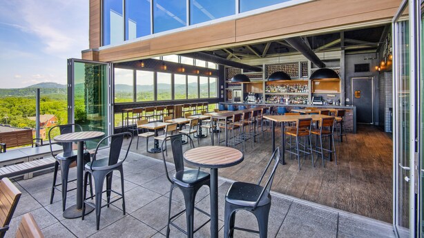 An outdoor dining area with multiple tables and chairs, a bar area with shelves of bottles, and large windows offering a view of the landscape. - DoubleTree by Hilton Asheville Downtown (Asheville)