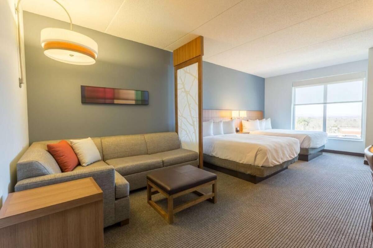 Una habitación de hotel con sofá, cama, mesita de noche, lámpara y ventana.