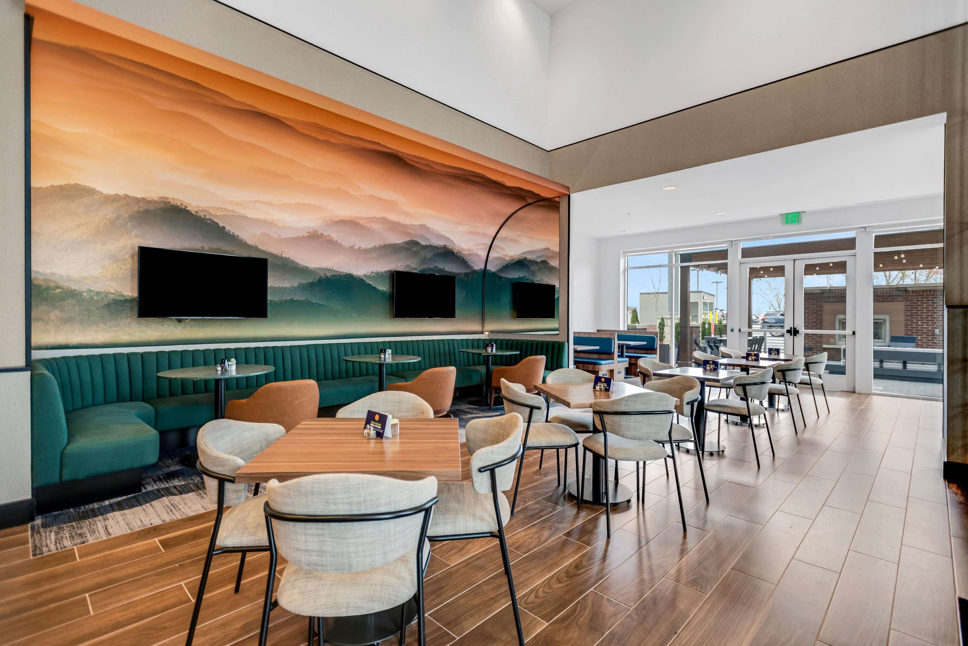 A modern restaurant with wooden tables and chairs, a green cushioned seating area, and a large mural on the wall.