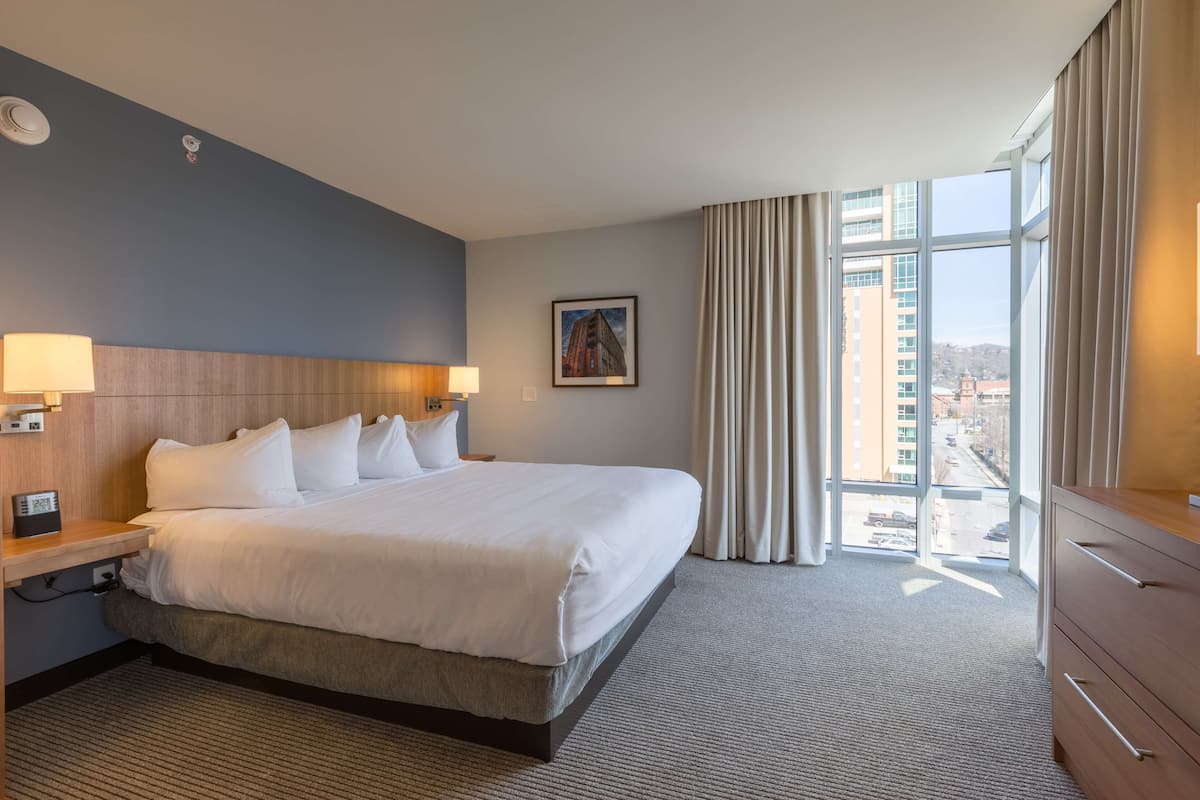 Ein Hotelzimmer mit einem großen Bett, einem hölzernen Kopfteil, einem Nachttisch mit Lampe, einem Schminktisch und einem Fenster mit Vorhängen.