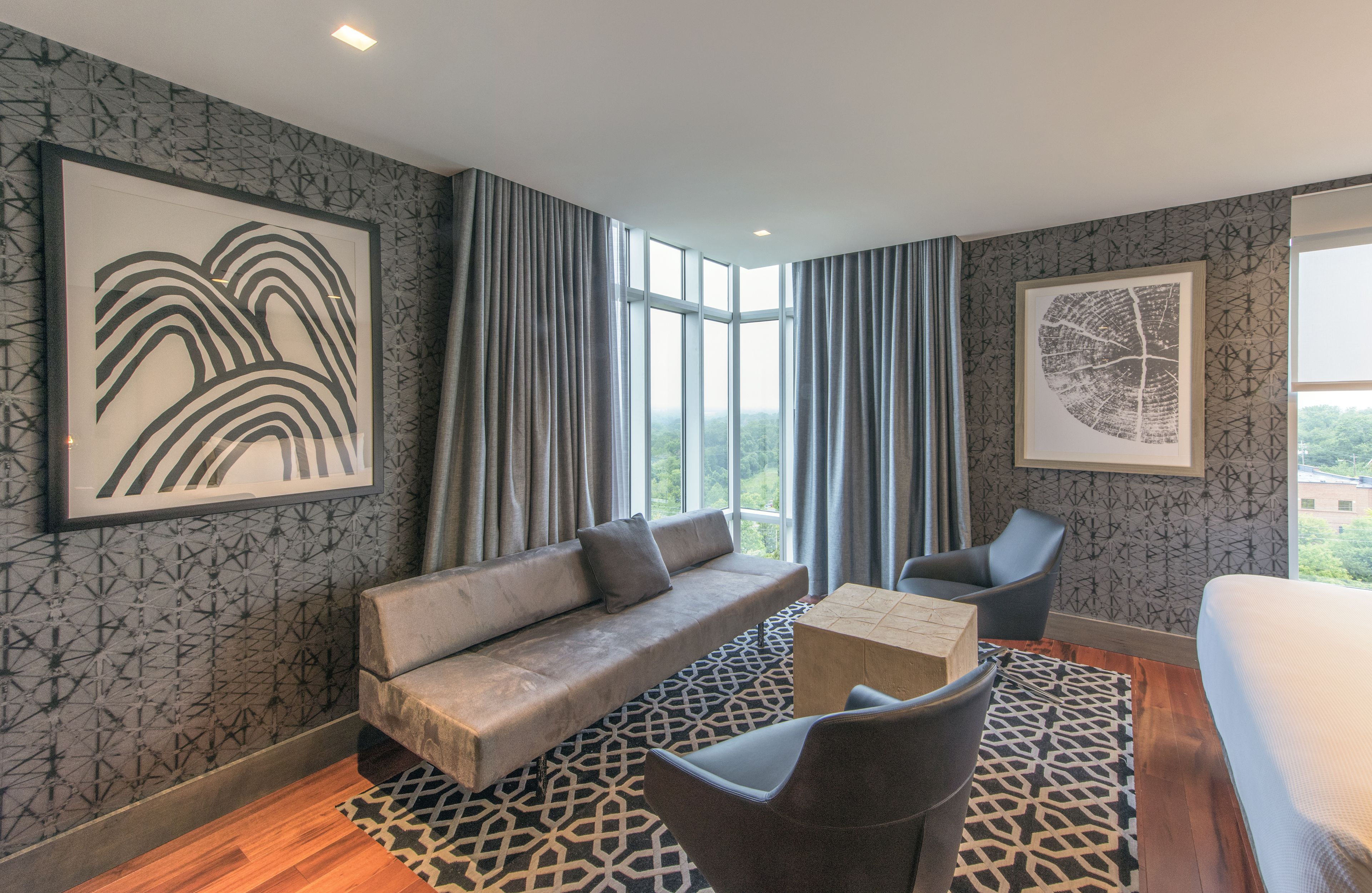A modern hotel room with a sofa, armchair, and a small table. There are large windows with curtains, a patterned wall, and a framed artwork.