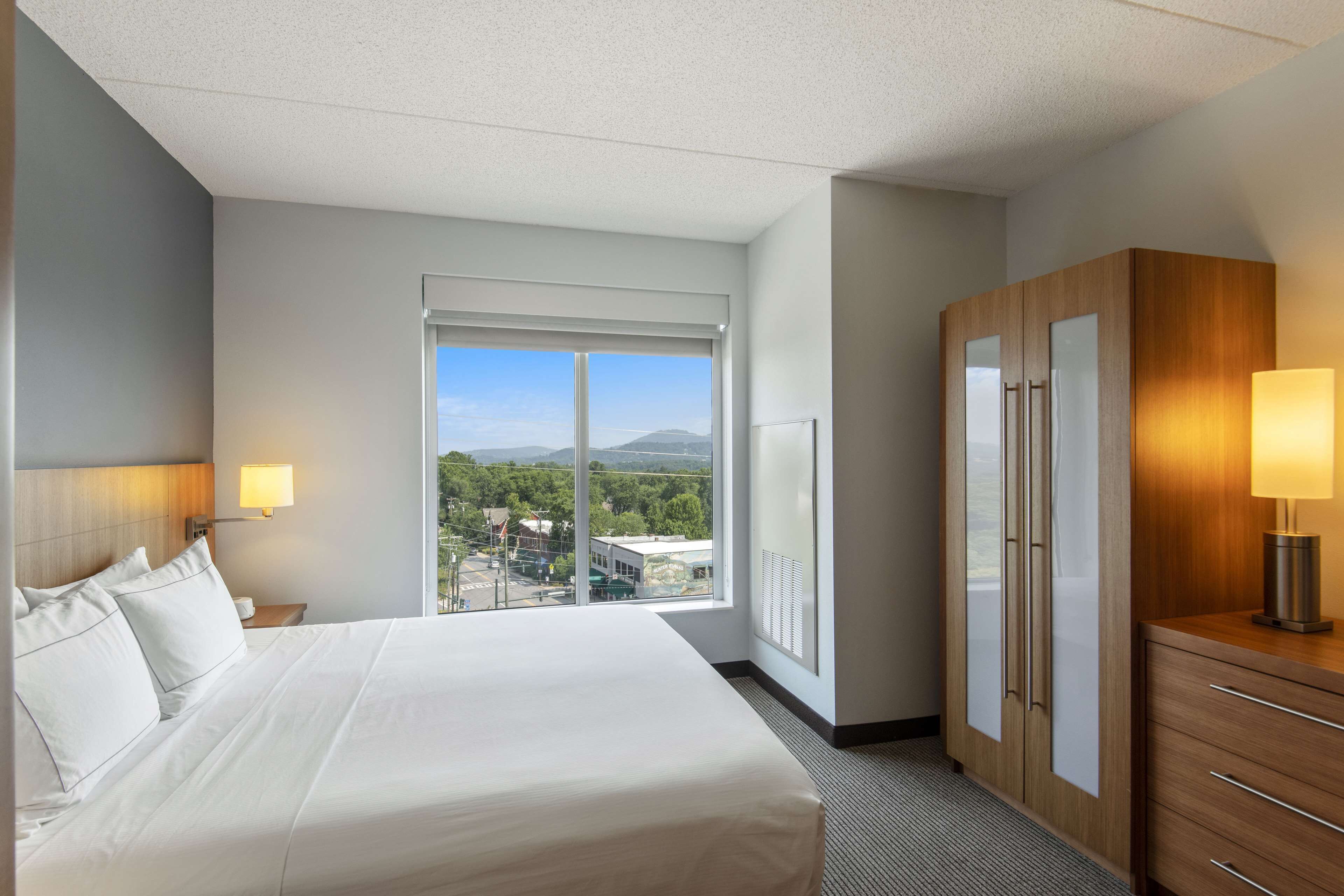 Una habitación de hotel con una cama grande, un armario, un escritorio y una ventana con vistas a la ciudad y a las montañas.