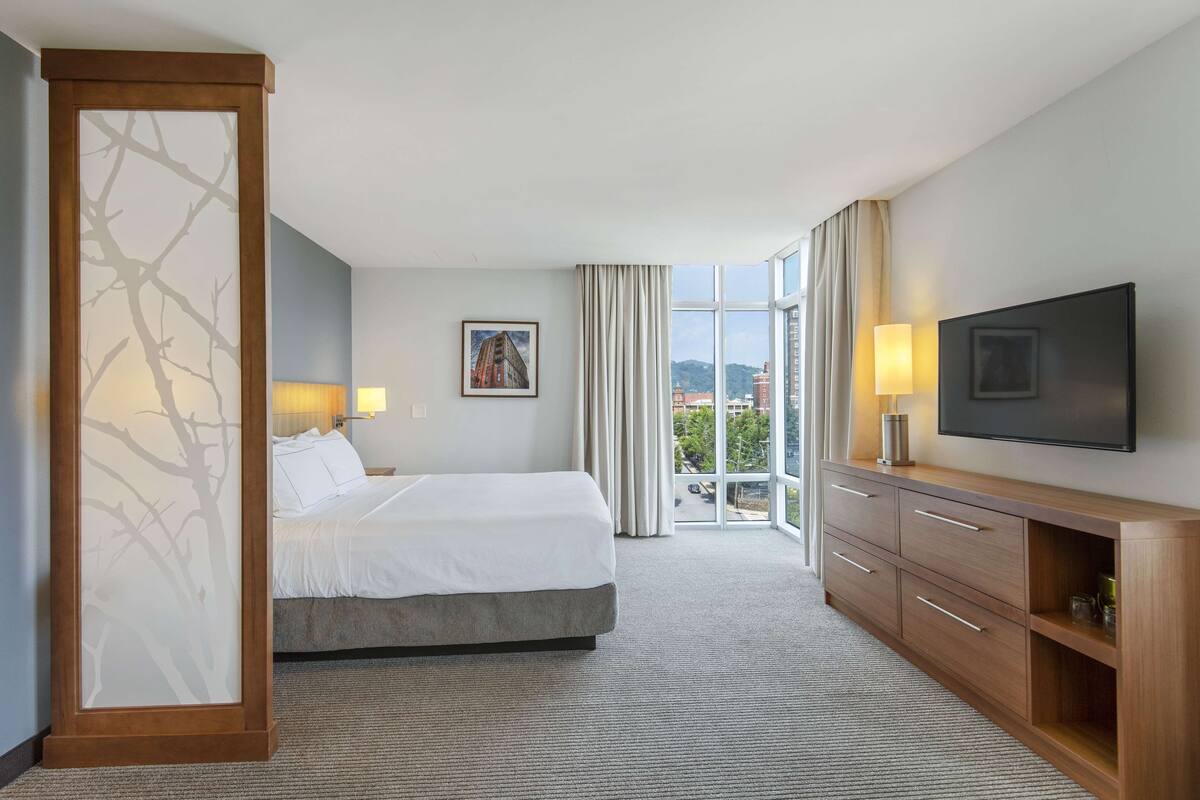 Ein Hotelzimmer mit Bett, Fernseher, einer Holzkommode, einer Lampe und Blick auf die Stadt durch große Fenster.