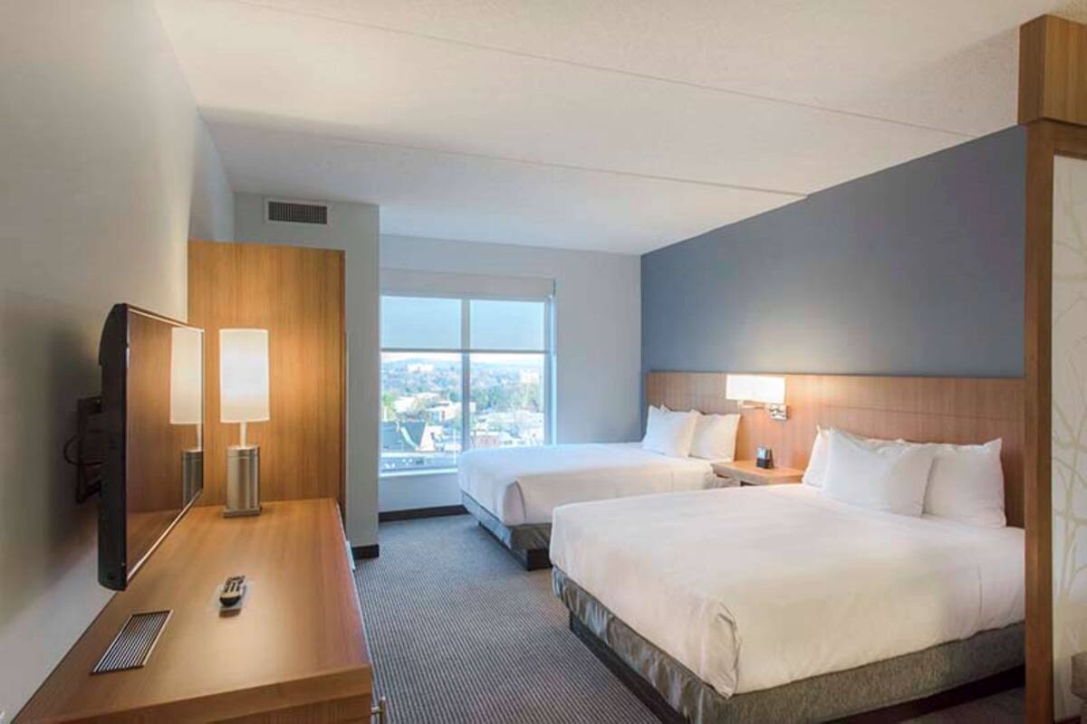 Ein Hotelzimmer mit zwei Betten, einem Schreibtisch mit Lampe, einem Fernseher und einem großen Fenster mit Blick auf die Stadt.