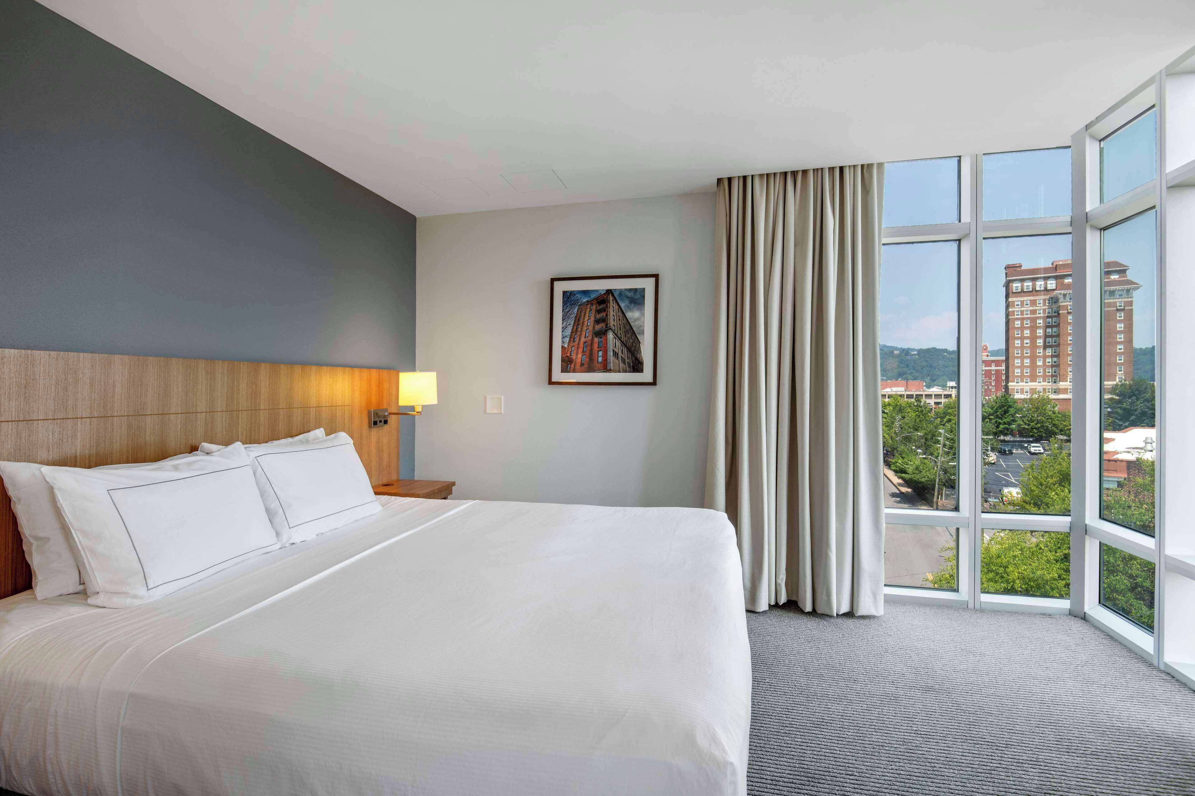 A hotel room with a large bed, a nightstand, a lamp, a framed picture on the wall, and a view of buildings through large windows.