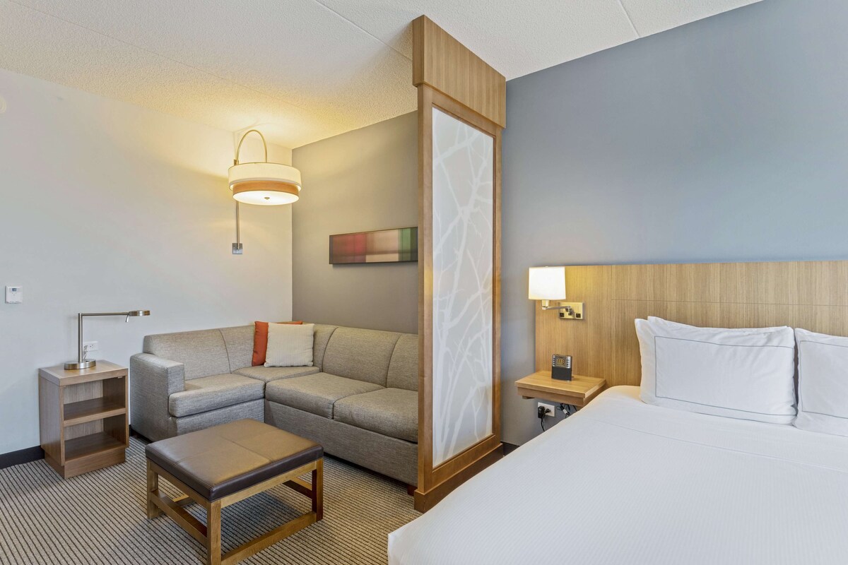 A hotel room with a bed, a sofa, a small table, a nightstand, a lamp, and a wall-mounted artwork.