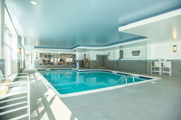 An indoor swimming pool with clear blue water, surrounded by white tiled flooring and walls, and equipped with a diving board and lounge chairs.