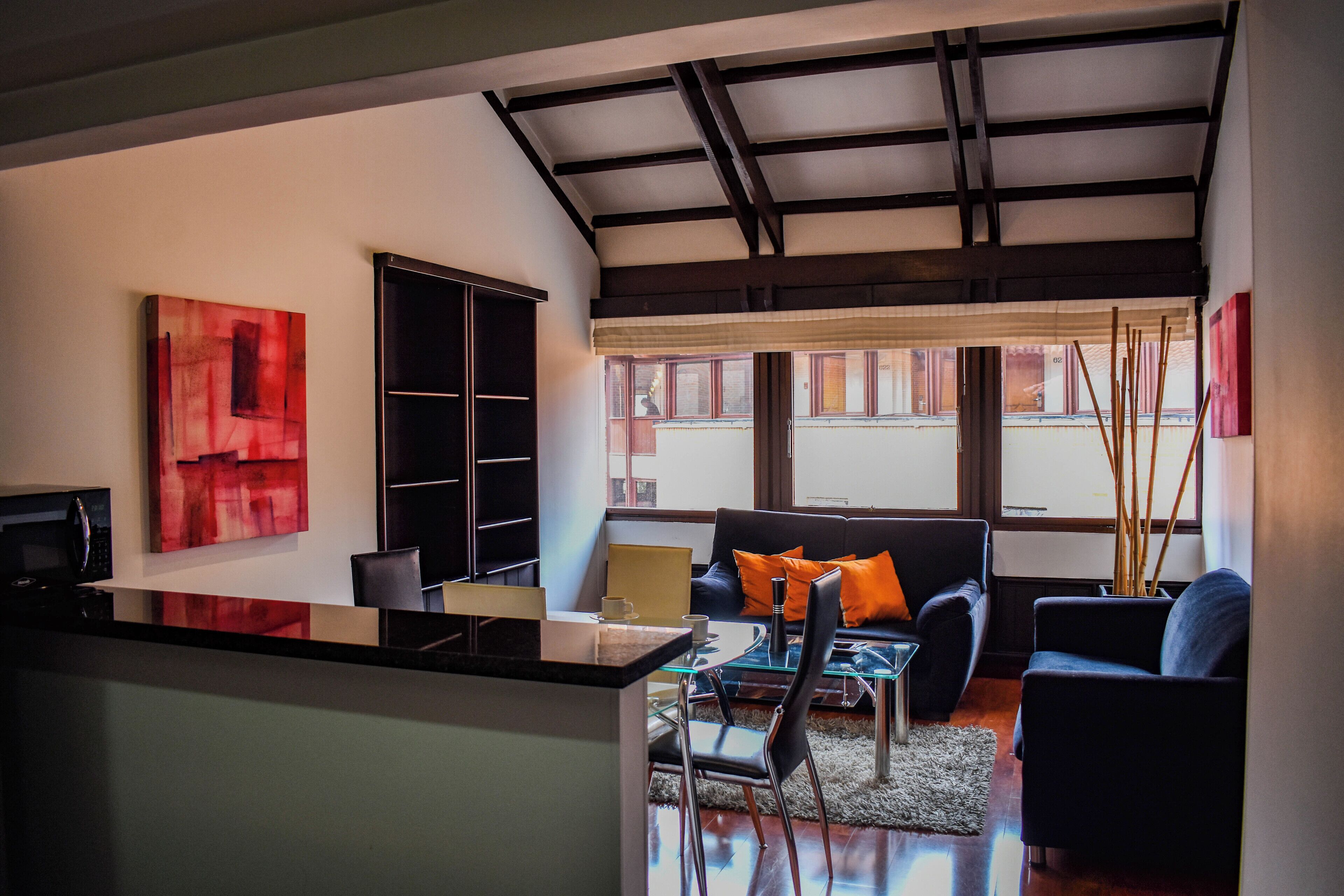 A modern living room with a black sofa, a glass coffee table, and a dining area with a black table and chairs. There is a large abstract painting on the wall and a black cabinet.