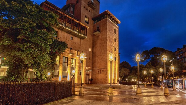 Un edificio histórico con una torre destacada y varios balcones, rodeado de árboles e iluminado por farolas.