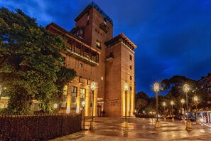 Un edificio histórico con una torre destacada y varios balcones, rodeado de árboles e iluminado por farolas.