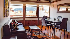 Una sala de estar moderna con un sofá, una mesa de centro y un comedor con sillas y una mesa. Amplias ventanas ofrecen vistas a edificios y montañas.