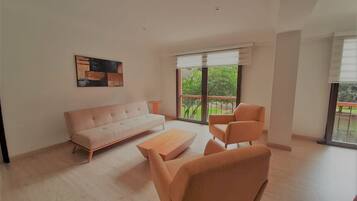 A spacious living room with a light-colored sofa, a coffee table, and a chair. There is a large window providing a view of greenery outside.