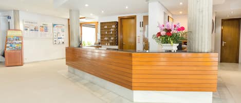 A reception desk with a wooden finish, a white base, and a large pillar to the right. There is a bulletin board with information and a flower arrangement on the desk.