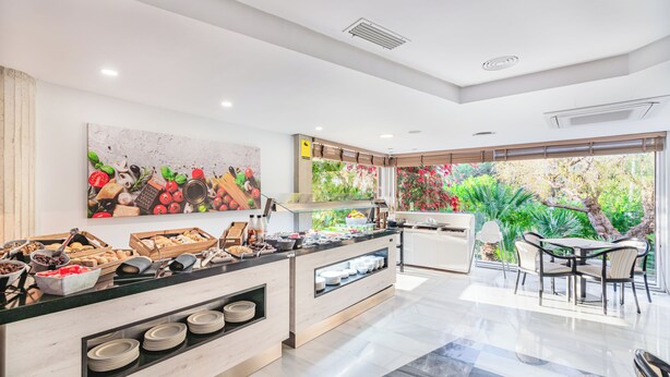 A modern hotel room with a breakfast bar, a large fruit and vegetable painting, a view of greenery, and a dining area with chairs.