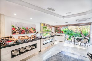 Una habitación de hotel moderna con desayunador, un cuadro grande de frutas y verduras, vista a follaje y un área de comedor con sillas.