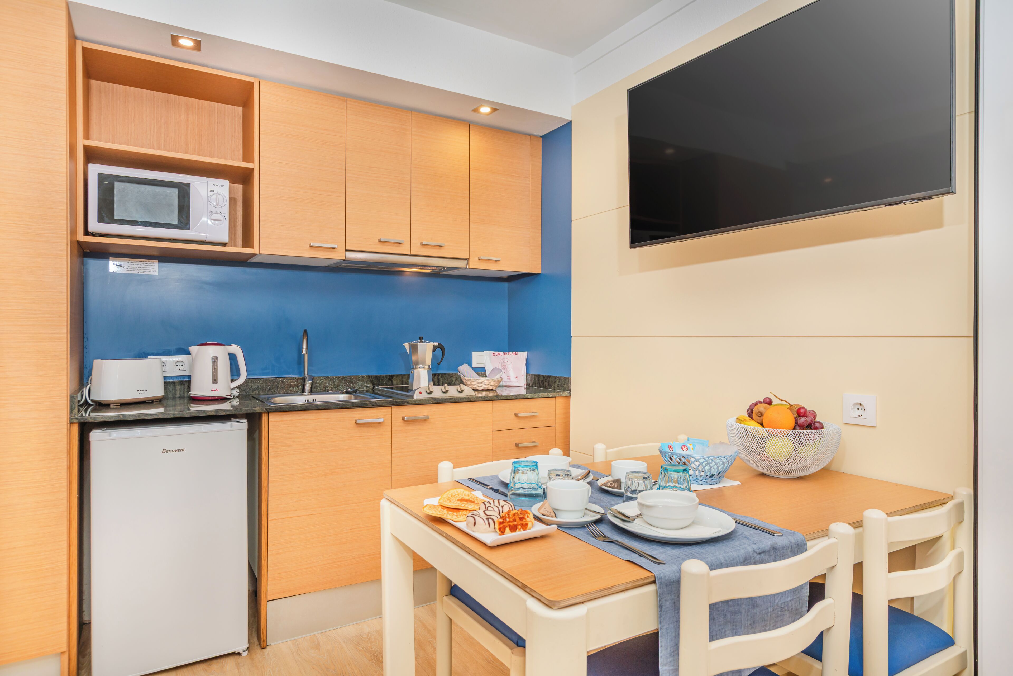 Uma cozinha compacta com micro-ondas, cafeteira e um pequeno frigorífico. Uma mesa de jantar com cadeiras está posta com artigos de pequeno-almoço.
