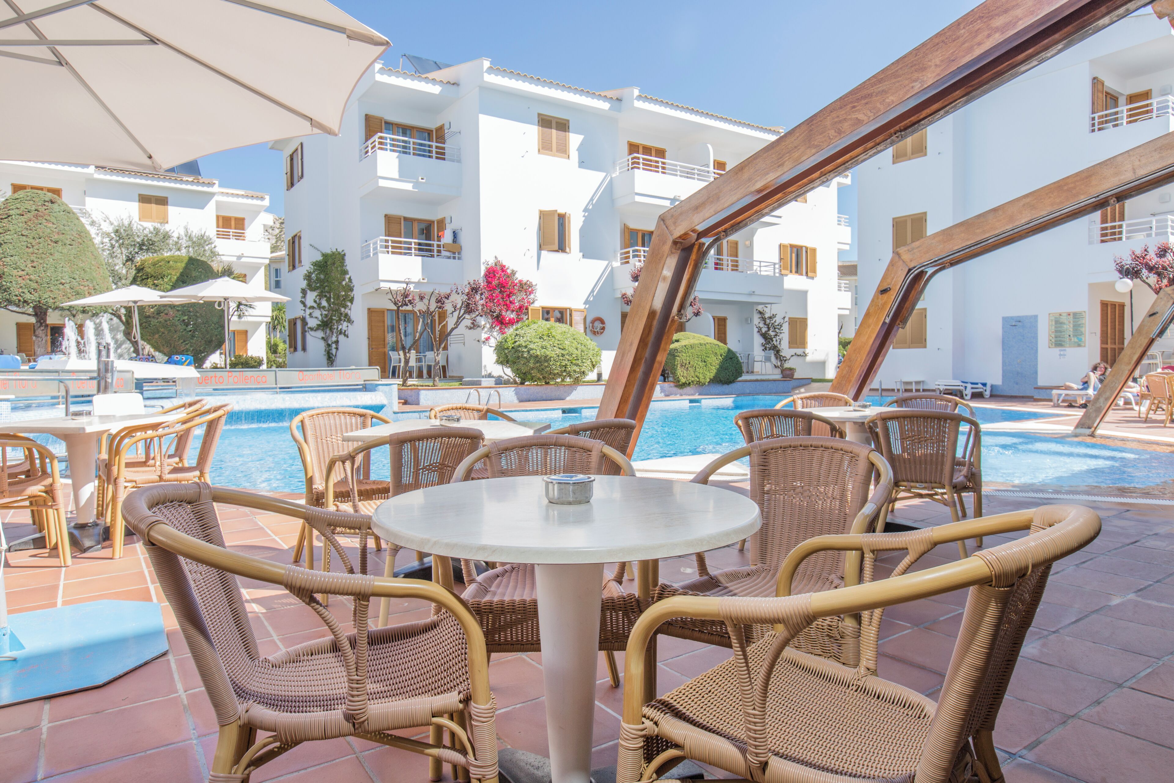 Zona junto a la piscina con mobiliario de mimbre, piscina y un edificio con balcones.