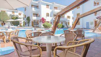 Zona junto a la piscina con mobiliario de mimbre, piscina y un edificio con balcones.