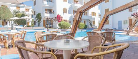 Zona junto a la piscina con mobiliario de mimbre, piscina y un edificio con balcones.