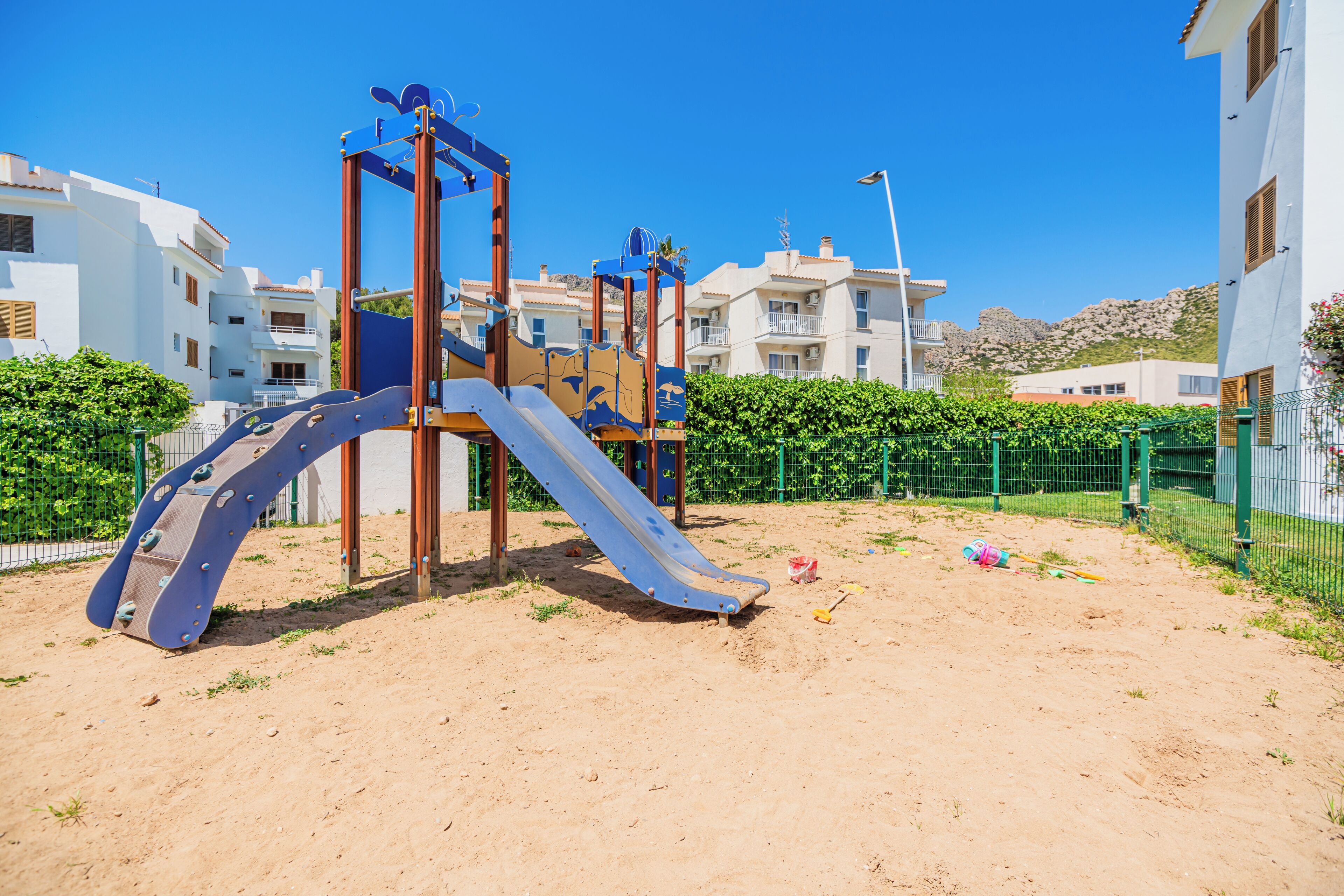 A playground with a slide and climbing structure, surrounded by a fence and buildings in the background.