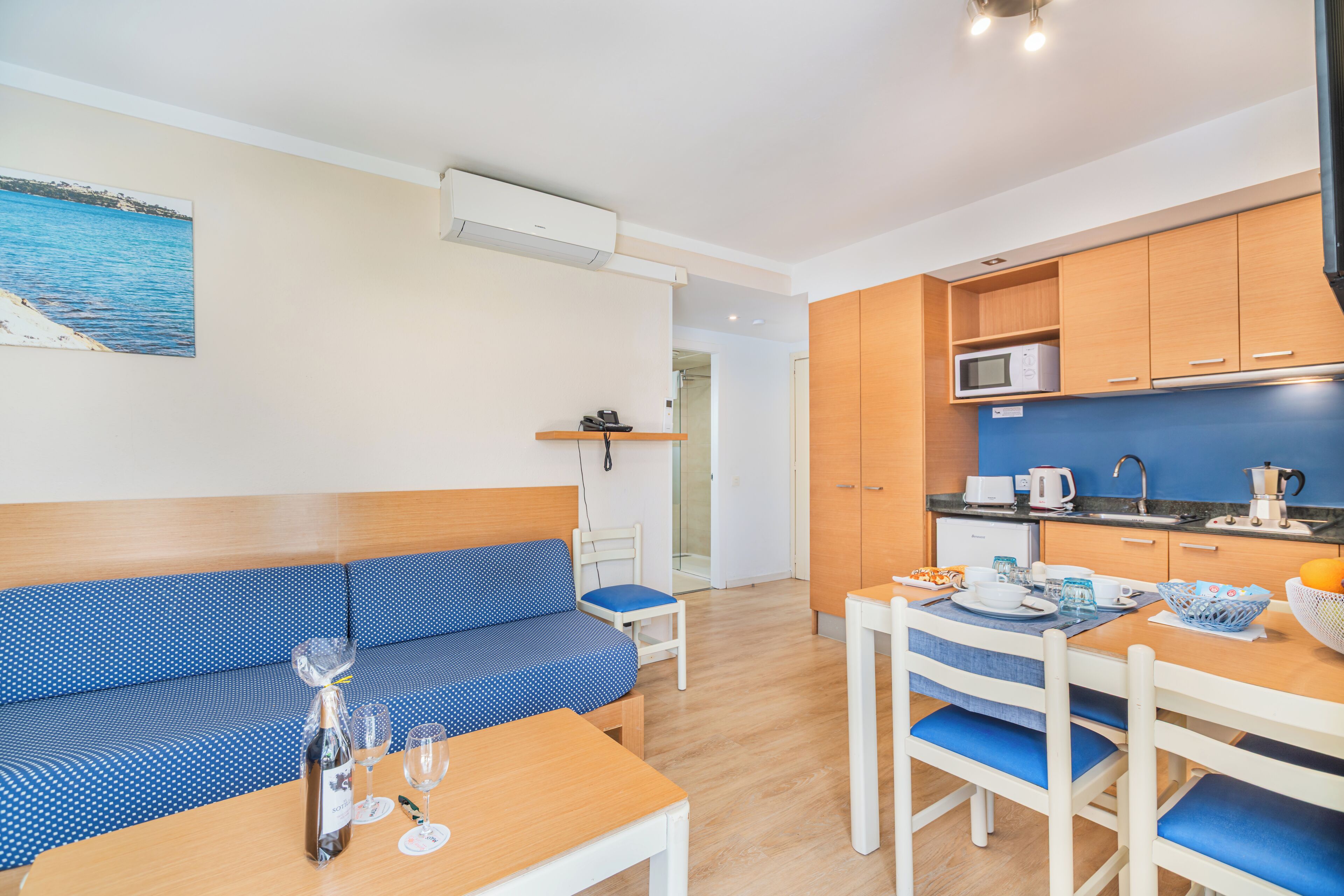Een moderne kamer met een bank, eettafel, stoelen, een keuken met kastjes en apparatuur, en uitzicht op zee door een raam.