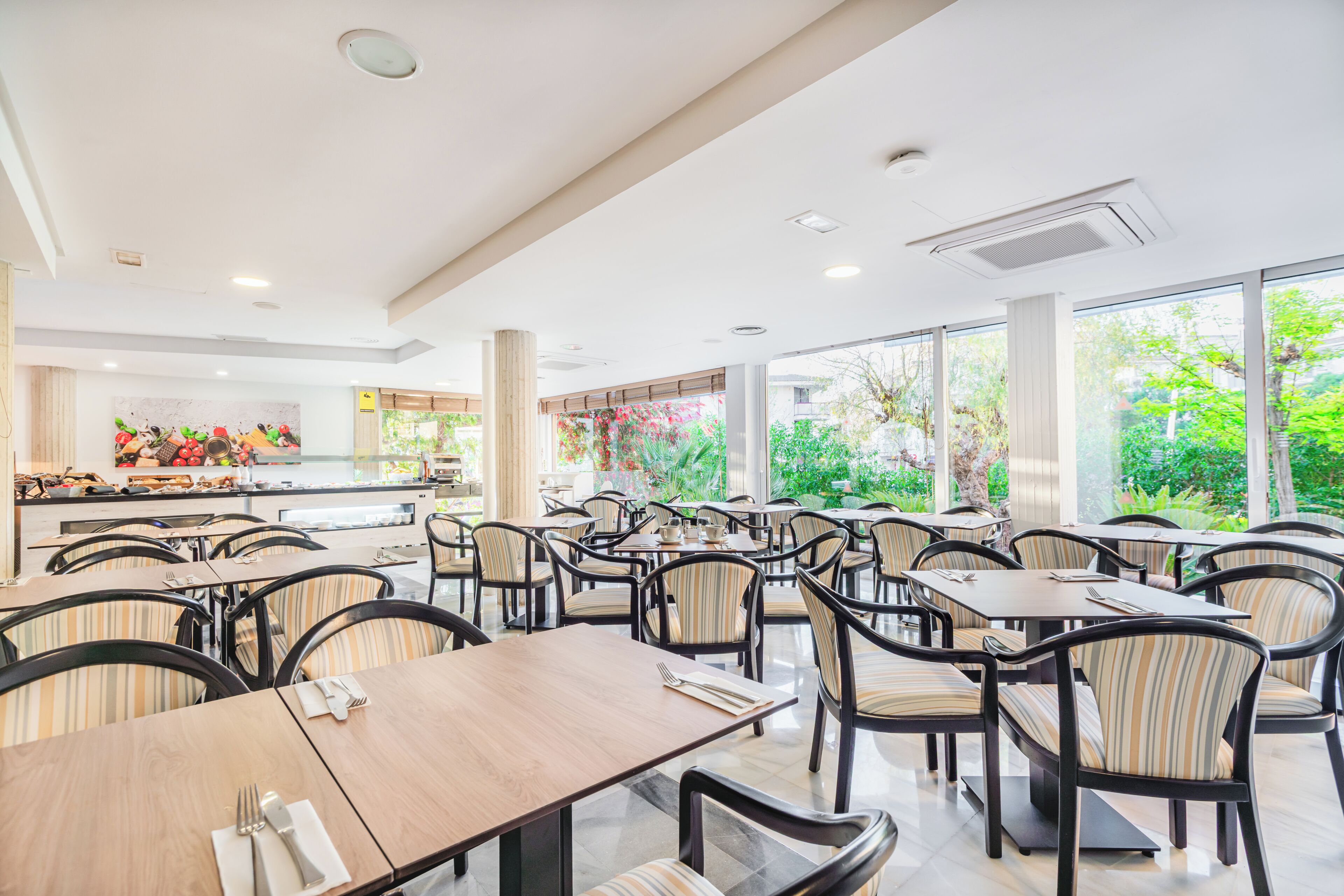 A spacious indoor dining area with multiple tables and chairs, a kitchen in the background, and large windows offering a view of greenery.