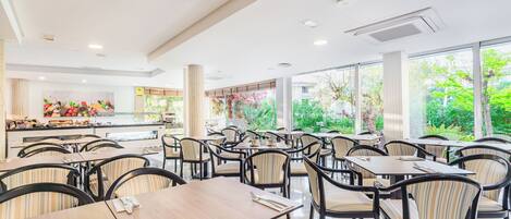 A spacious indoor dining area with multiple tables and chairs, a kitchen in the background, and large windows offering a view of greenery.
