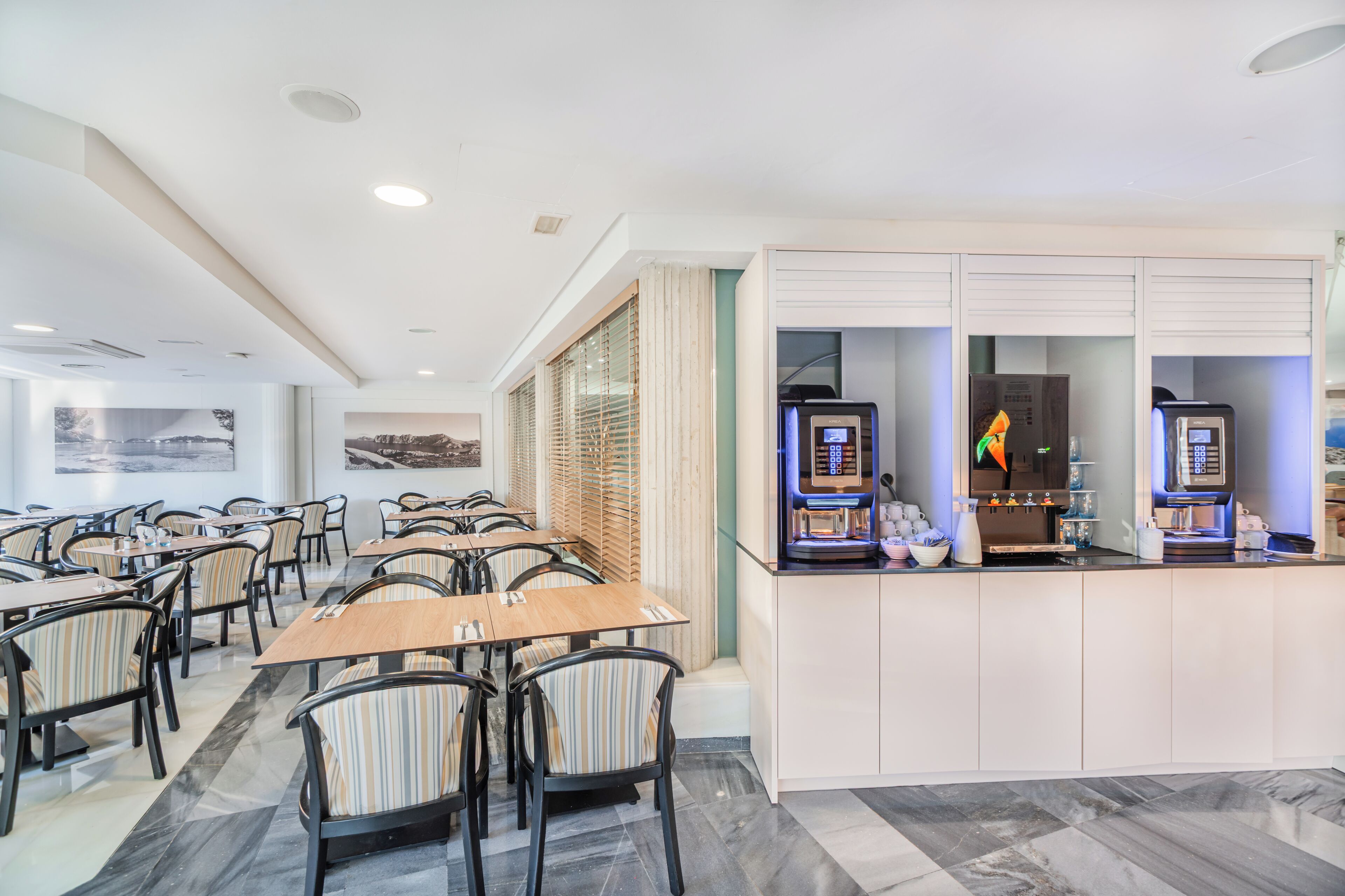 Een restaurantgedeelte in een hotel met tafels, stoelen, een koffieapparaat en uitzicht op de stad.