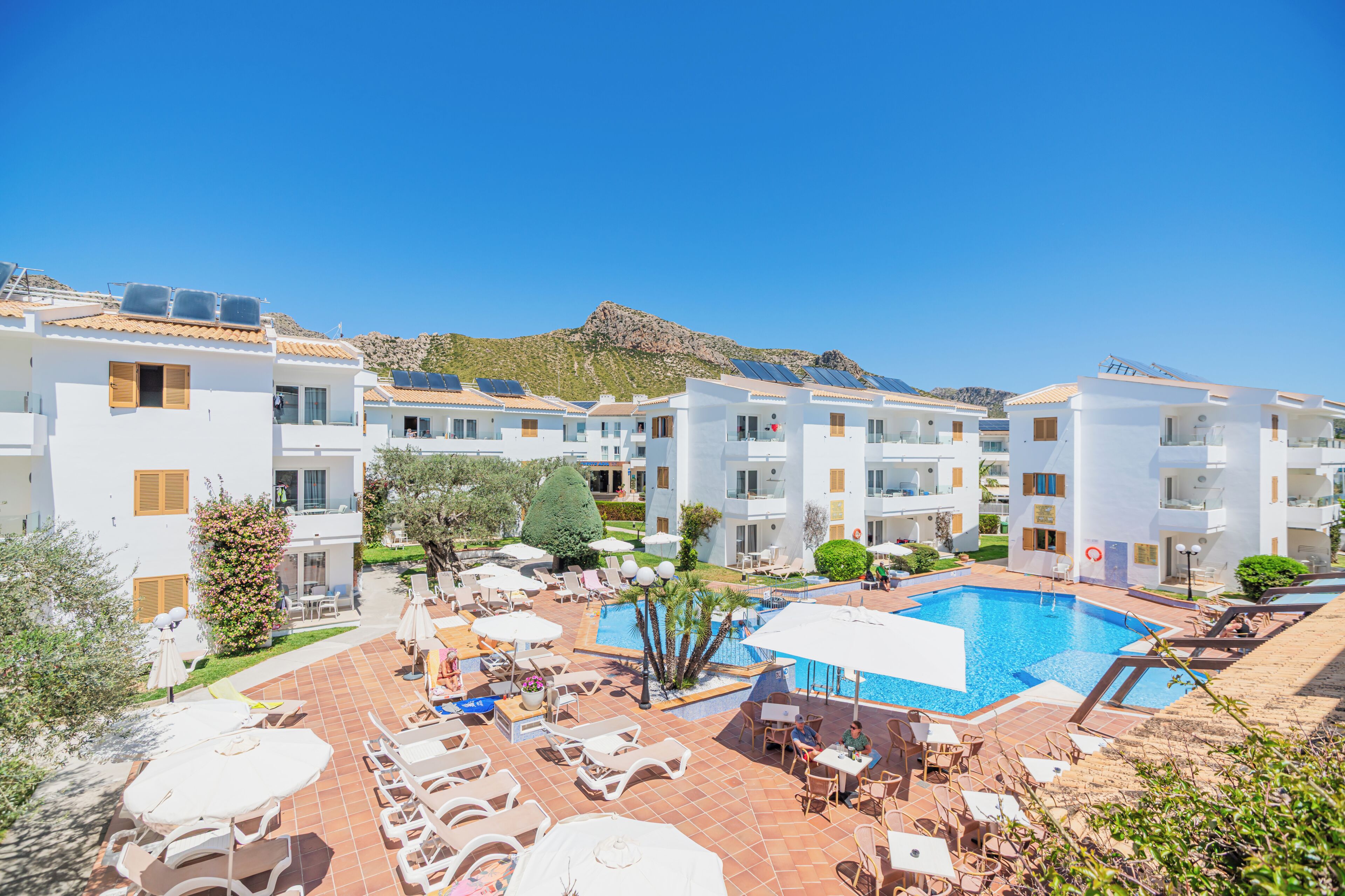 Ein Resort mit mehreren weißen Gebäuden, einem Swimmingpool, Liegestühlen und einem Berg im Hintergrund.