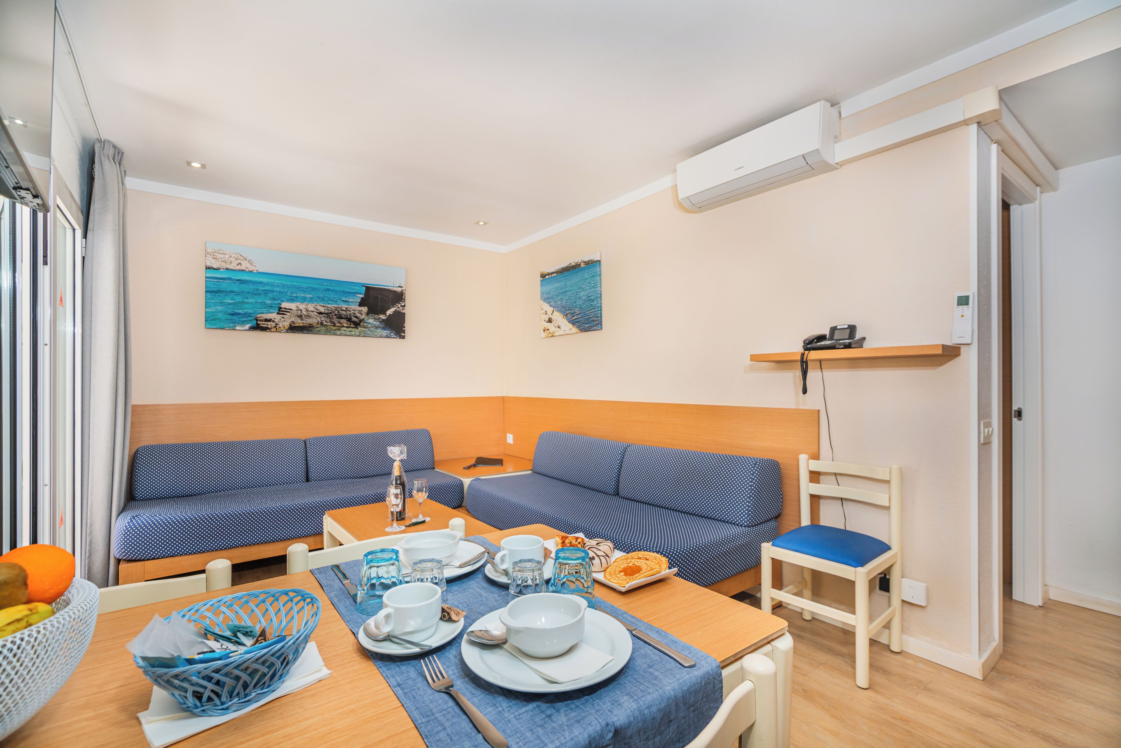 A compact living space with a dining area, two blue sofas, a small table set for two, and a wall-mounted shelf with a phone.