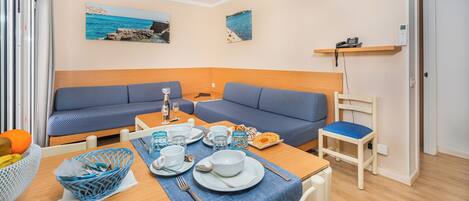 A compact living space with a dining area, two blue sofas, a small table set for two, and a wall-mounted shelf with a phone.