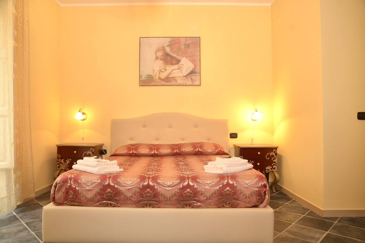 A neatly made bed with a patterned bedspread and two pillows, flanked by nightstands with lamps, in a room with a warm yellow wall and a framed artwork above the bed.