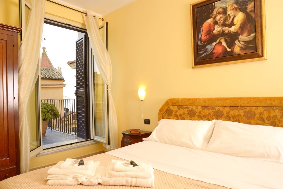 A hotel room with a bed, a window with curtains, a painting on the wall, and a nightstand with a lamp.